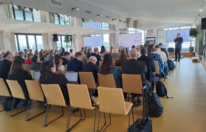 An audience seated in rows listens to a presentation in a bright conference room, with presentation slides displayed on several screens.